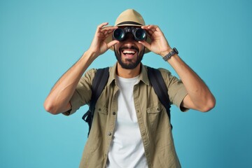 Smiling man with binoculars on blue background, ready for adventure, wearing backpack and casual outdoor clothing