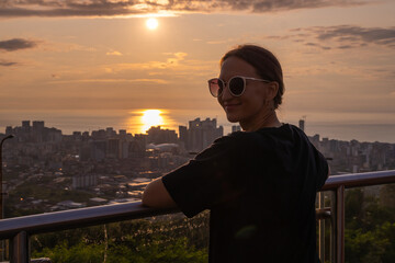 Fototapeta premium Young woman is leaning on a railing, enjoying a beautiful sunset over the cityscape. Sun is reflecting on the sea creating a romantic atmosphere