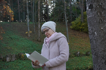 Positive senior woman with gray hair reading book in an autumn park.