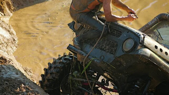 The mud-covered co-driver of racing team is sitting on the hood of an off-road vehicle. A man is holding a winch cable. The front of the SUV is on a steep slope and the back of is in a deep puddle