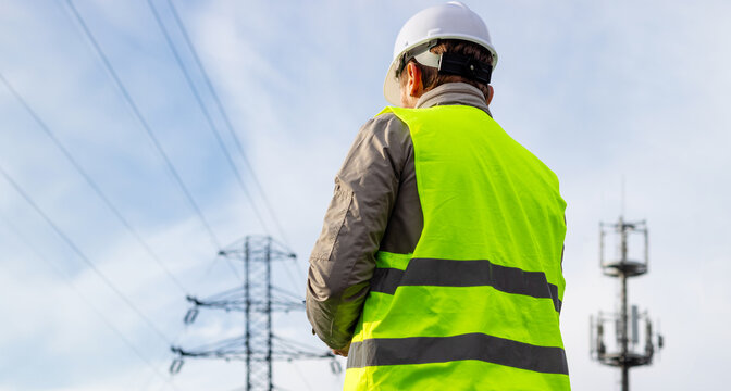 Male engineer, seen from the back, employee of a telecommunications company, stands in front of a high voltage power line and a cell phone tower.