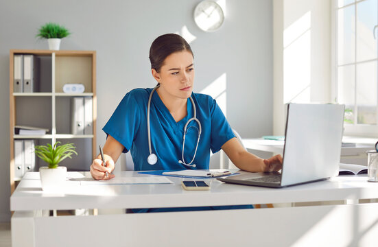 Portrait of a young female doctor or nurse in blue medical uniform sitting at the desk and working in office. Serious woman physician wearing stethoscope in clinic using laptop and typing.