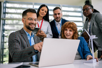 Multiracial business people talking about sustainable energy. They have windmill on a table and they are doing something on a laptop.