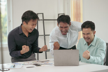 Three euphoric businessmen are raising their fists in the air while looking at a laptop, celebrating achieving business goals