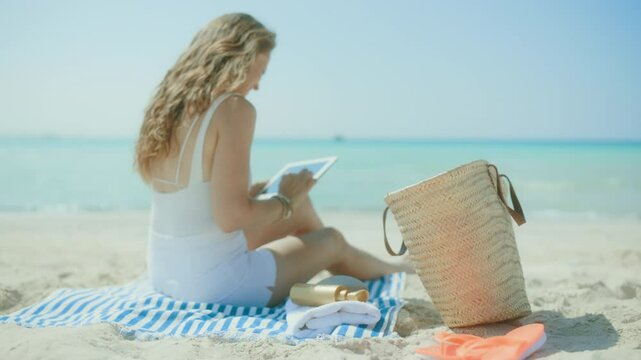 relaxed modern middle aged woman on the ocean shore with straw bag and striped towel using applications on tablet PC.