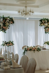 A wedding reception with a white backdrop and floral decorations. The room is filled with white chairs and tables