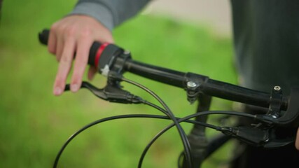 Close-up of individual wearing black trousers and grey cloth, approaches parked bicycle, adjusting brake lever in grassy field, the person holds the handlebar with firm grip