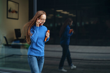 Casual woman in a blue sweater talking on the phone while walking past a modern office building on a sunny day