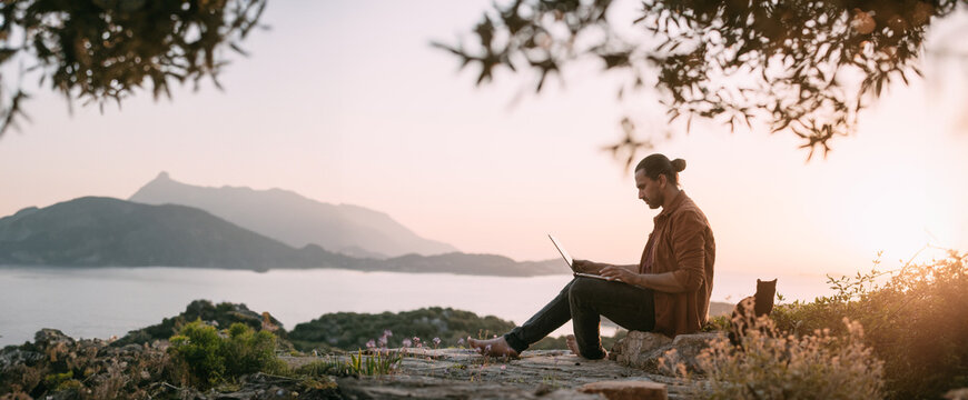 A Caucasian man is working with a laptop in a garden on a mountain overlooking the sea and sunset.