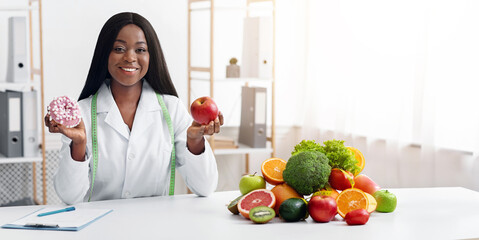 Pretty dietary doc holding donut and apple, posing at her office, healthy food concept