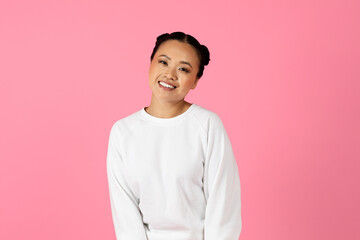 A cheerful young Asian woman stands confidently with a bright smile, wearing a simple white sweater. The vibrant pink backdrop enhances her joyful expression, creating a lively atmosphere.