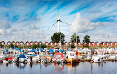 A vibrant marina in sweden filled with boats, framed by a row of red cabins and a large wind turbine in the background, under a clear blue sky.