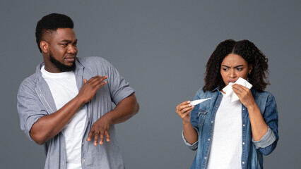 Unpleased Black Man Avoid His Ill Girlfriend That Standing With Thermometer In Hands Over Light Studio Background, Checking Temperature