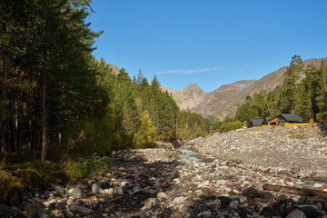 Autumn mountain landscape. A gorge with a rocky riverbed and a chalet in a pine grove. In the distance on the mountainside cable car.
