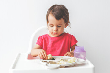 girl child in a bright pink t-shirt sitting in a high chair on a white background and having lunch, baby food concept