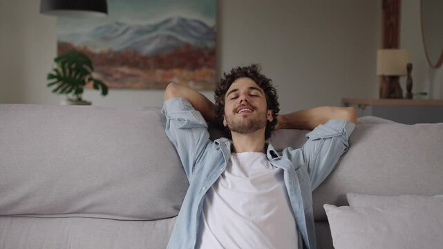 Happy relaxed young man rest lounge lean on couch enjoy peaceful mood, healthy lazy calm guy dreaming breathing fresh air sit on comfortable sofa in living room on stress free cozy day at home

