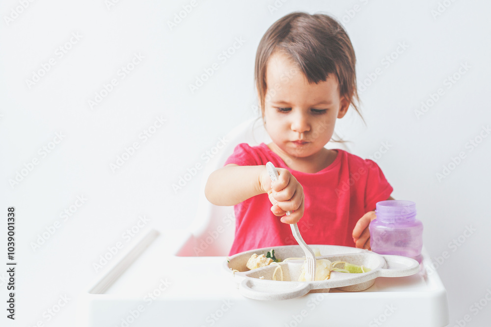 Wall mural cute 1.5 year old girl sits in a high chair and learns to eat with a fork, concept of childhood and 