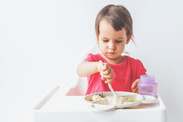 cute 1.5 year old girl sits in a high chair and learns to eat with a fork, concept of childhood and baby food.
