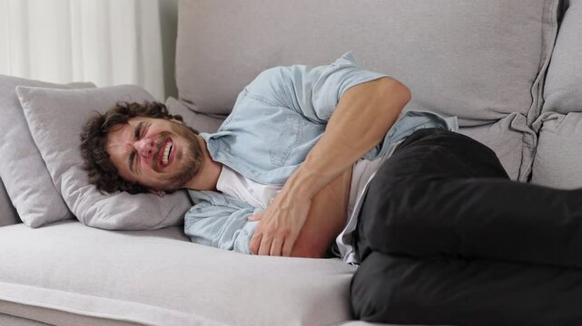 Terrible pain in the stomach. A young man suffering from abdominal pain, holding his stomach, lies on the sofa
