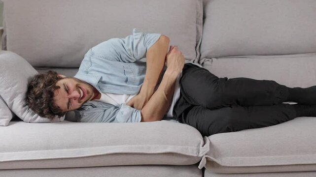 Young caucasian man lying on sofa feeling sudden strong abdominal stomach ache, gastritis problem. White guy having symptom poisoning diarrhea indigestion peptic ulcer pancreatitis at home apartment r