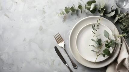 Top view of a table setting. White plate, cutlery and green leaves on gray background