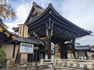 Kyoto, Japan - 2023.12.14: Hongan-ji Goeidomon, the main entrance to the Nishi Hongan-ji featuring wooden structure and decorated rooftop with tiles next to a stone bridge under a blue sky with clouds