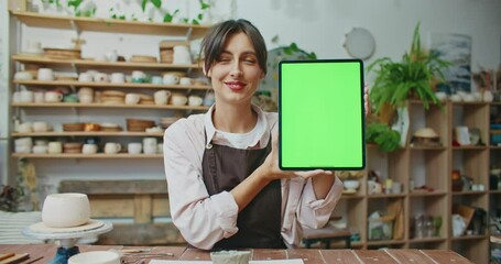 Cute Caucasian girl looking at tablet screen before glancing at camera and smiling. Chroman key visible on large display. Green screen. In background many shelves with finished tableware.