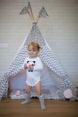 Adorable beautiful happy smiling ginger-haired toddler little girl is playing with her toys and drinking tea standing at the teepee wigwam © Tina