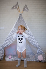 Adorable beautiful happy smiling ginger-haired toddler little girl is playing with her toys and drinking tea standing at the teepee wigwam © Tina