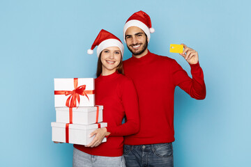 A cheerful couple dressed in festive red sweaters and Santa hats smiles while holding wrapped Christmas gifts. The bright blue background enhances the holiday spirit.