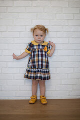 Adorable beautiful happy ginger-haired toddler baby girl in a checkered yellow dress in autumn style and yellow shoes stands at a white wall