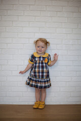 Adorable beautiful happy ginger-haired toddler baby girl in a checkered yellow dress in autumn style and yellow shoes stands at a white wall