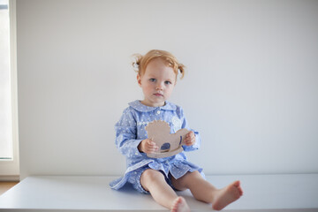 Aesthetic minimalistic portrait of Adorable beautiful smiling happy ginger-haired toddler baby girl sits on a white chest of drawers and plays with a toy horse