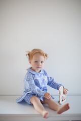 Aesthetic minimalistic portrait of Adorable beautiful smiling happy ginger-haired toddler baby girl sits on a white chest of drawers and plays with a toy horse