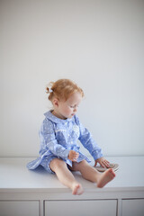 Aesthetic minimalistic portrait of Adorable beautiful smiling happy ginger-haired toddler baby girl sits on a white chest of drawers and plays with a toy horse