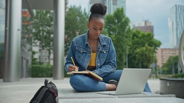 Concentrated African American female sitting next to staircase in front of modern building. Student looking at laptop screen. Writing down information with pencil in notebook. Studying additionally.