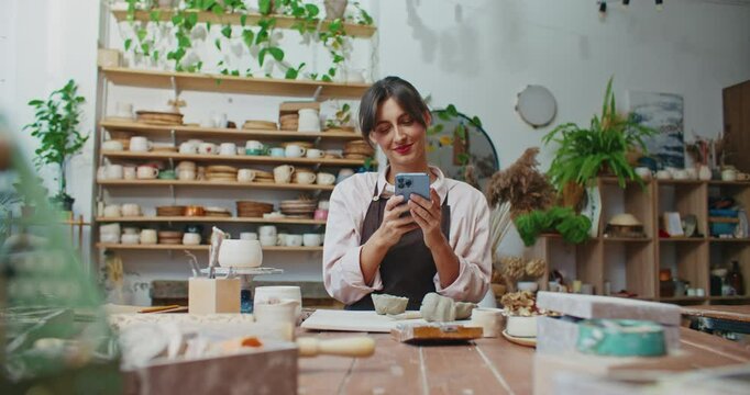 Cute Caucasian girl typing message on smartphone. Finishing typing text on screen with fingers. Placing mobile phone down. Continuing working as potter. Wedging and Kneading piece of clay.