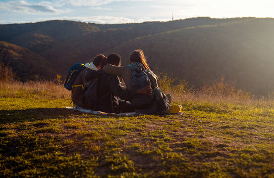 Rear view group of friends sitting on the top of the hill resting after hiking and looking towards the sunset.