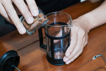 Close up of a person's hands pouring coffee powder in a black french press on the wooden table, Brewing a Coffee in a French Press at home.