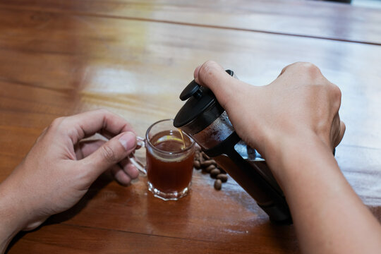 Closeup of a hand man pouring coffee from French press coffee maker into a small glass on the wooden table, making coffee homemade.