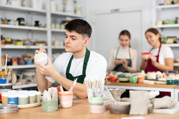 Concentrated young man in apron engraving on clay cup with cutter in pottery workshop