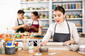 Fototapeta premium Interested young woman in pinafore rolling row clay with rolling-pin in art studio. Persons viewing handmade pottery products