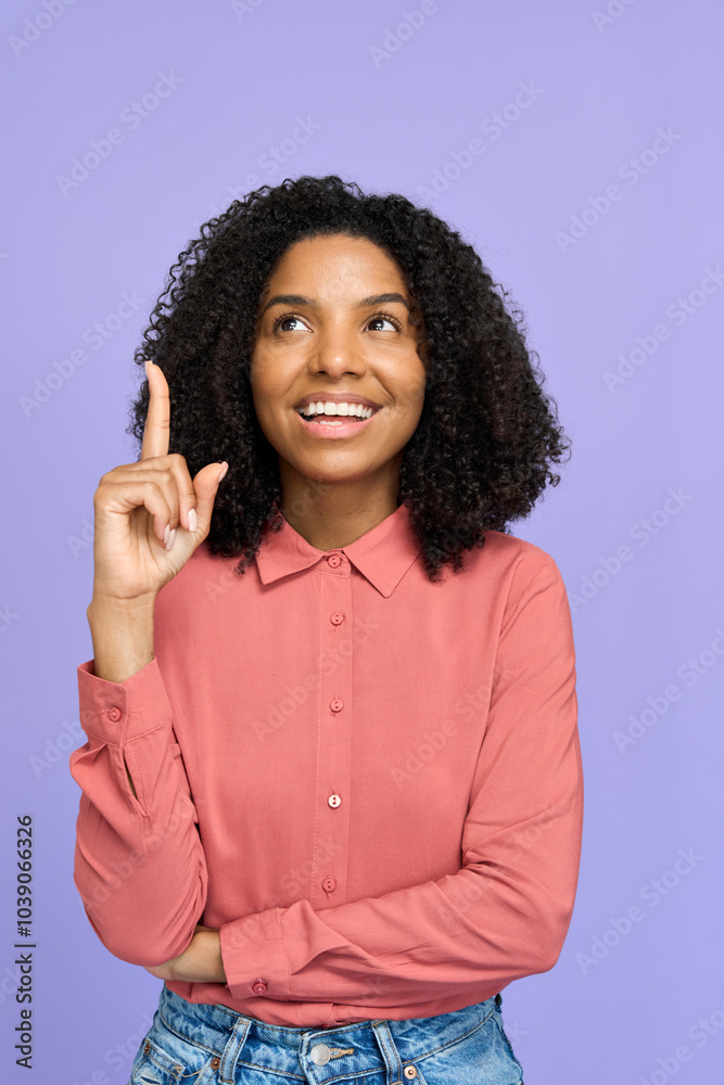 Wall mural Happy young African woman standing isolated on purple background presenting promotion ads. Smiling lady wearing pink shirt looking upward pointing up at copy space, having new idea.