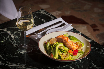 A plate of grilled salmon, avocado and salad with a glass of white wine. Luxurious and expensive gourmet meal on a black marble table.