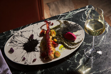 Octopus dish prepared like a work of art. Octopus dish garnished with grilled vegetables and special sauces. Glass of white wine on black marble table. Aphrodisiac dishes, close-up