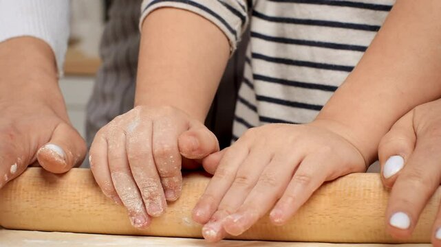 A parent and child work together, pressing their hands on a rolling pin to flatten dough while surrounded by flour in a warm kitchen.