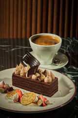Chocolate cake and a cup of coffee. Cake decorated with drops of cream and chocolate paper, next to it is a dessert plate prepared with edible decorations such as strawberries, figs and oranges.