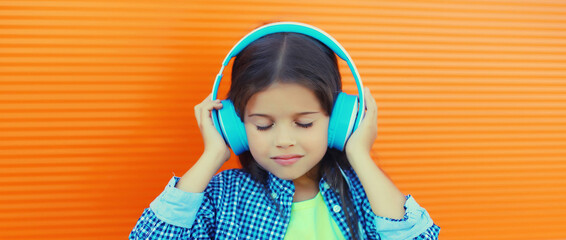 Happy little girl child in blue wireless headphones listening to music on orange background