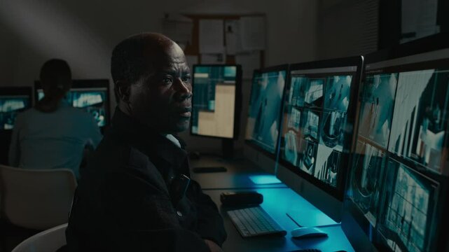 Medium closeup side shot of black male security guard sitting at desk in dark control room, observing CCTV footage of people walking around business premises, watching for abnormal behaviour