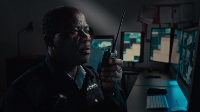 Tilting closeup of hands and face of frowning middle-aged African American security guard watching CCTV camera footage, then picking up radio, calling colleague to report abnormal customer behaviour